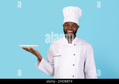 Ritratto di giovane giovane cuoco nero sorridente in uniforme che tiene piatto vuoto Foto Stock