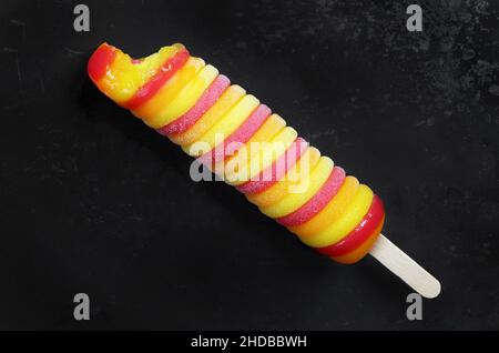 Bitten colorato gelato di frutta ritorto su un bastone su vecchio sfondo di metallo nero da vicino, vista dall'alto Foto Stock