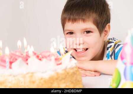 Un ragazzino ride, festeggia il suo compleanno, si siede di fronte ad una torta bianca con candele. Foto Stock