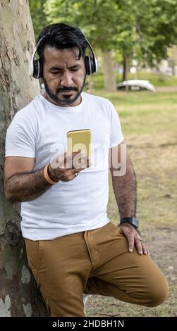 Latino uomo appoggiato contro un albero guardando il suo cellulare giallo Foto Stock