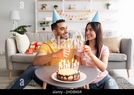 Coppia allegra e variegata in cappelli festosi tostando con bicchieri di champagne, avendo torta di compleanno con candele a casa Foto Stock