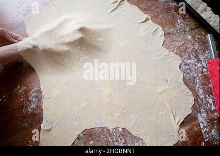 Vista dall'alto di una donna che tira e stretching pasta di pasticceria vegana fatta in casa su un tavolo da pranzo. Foto Stock