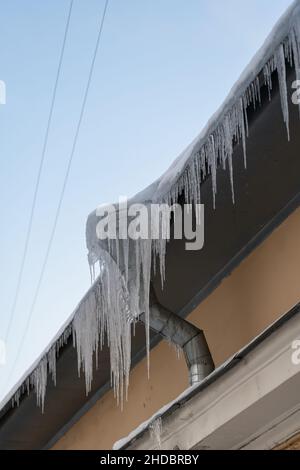 Grandi ghiacciate che pendono pericolosamente dal bordo della costruzione nella fredda giornata invernale. Diga di ghiaccio sul tetto Foto Stock