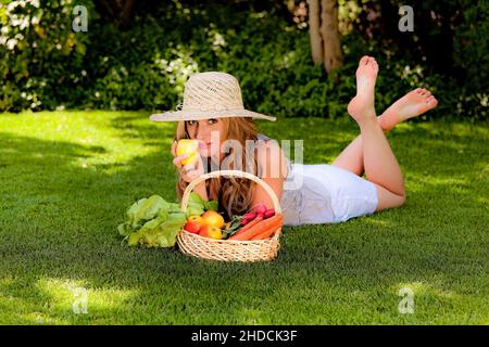 Blonde junge Frau liegt auf einer Wiese im Park und geniesst Obst und Gemüse, Picknick, Strohhut, Foto Stock