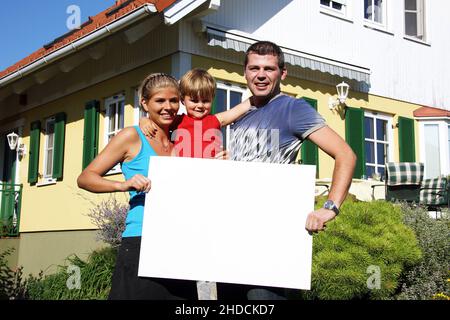 Junge Familie mit tipo neugekauftem vor Haus, hält weisses Schild, frei für beliebigen Testo, 30,35, 5 Jahre, signor:Sì Foto Stock