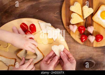 La famiglia prepara il cibo a forma di cuore, vista dall'alto. Salsicce, formaggi, sandwich a forma di cuore con pomodori ciliegini e toast. San Valentino. Foto Stock