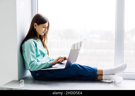 Ragazza carina seduta sul davanzale della finestra con il laptop che studia in linea a casa. Concetto di istruzione remota. Foto Stock