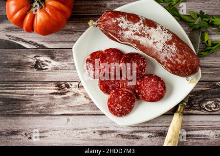 Vista aerea di Salami intero e affettato sottile su un piatto bianco su un rustico tavolo in legno. Rustico concetto di cibo di paese. Foto Stock