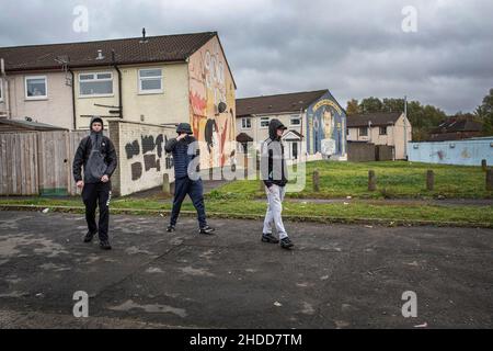 Giovani che camminano di fronte a murales politici con simboli paramilitari sulla Lower Shankill Estate, Belfast. Foto Stock