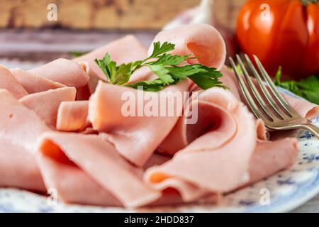 Primo piano di una disposizione rustica di un prosciutto squisito su un vecchio piatto con una forchetta e un pomodoro biologico sullo sfondo su un tavolo di campagna. Foto Stock