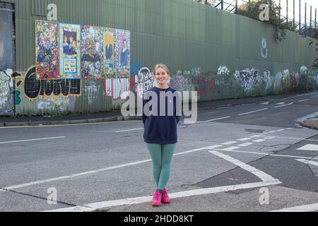 Amy Rafferty di Trademark, Community Organisation fuori dal suo ufficio con dipinti e murales colorati sul muro della Pace, o Peace Line, che corre lungo la Coppa Foto Stock