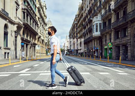 Giovane maschio con una maschera che porta una valigia che attraversa una strada Foto Stock
