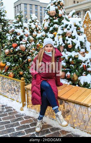 Una bella ragazza si siede su una panca vicino ad un albero di Natale in inverno. Natale, Capodanno. Foto Stock