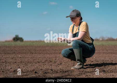 Agronomo agricoltore donna che controlla la qualità del terreno arato prima della stagione di semina, trattore agricolo in background, fuoco selettivo Foto Stock