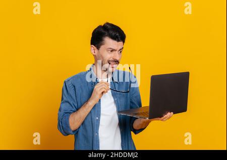 Scoraggiato dispiaciuto moderno caucasico in camicia denim, tiene un laptop, sembra confuso allo schermo, tenendo i suoi occhiali, senza umore, si erge su un isolato sfondo arancione Foto Stock