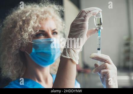 Una siringa che viene aspirata con il vaccino Covid-19. Primo piano verticale. Medico curly capelli femmina indossare maschera medica e guanti. Foto Stock