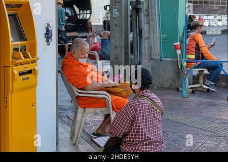 Un monaco buddista tailandese arancione seduto accanto a un bancomat giallo considera quali biglietti per la lotteria acquistare da un venditore di lotterie; Bangkok, Thailandia Foto Stock