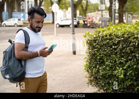 Uomo latino con barba che cammina in ambiente urbano guardando il suo cellulare Foto Stock