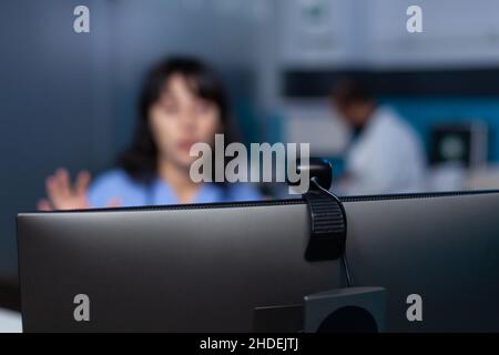 Primo piano della webcam sul computer utilizzato per le videochiamate e la comunicazione remota presso l'ufficio medico. Infermiera con telecamera sul monitor per videoconferenza online e telemedicina a tarda notte. Foto Stock