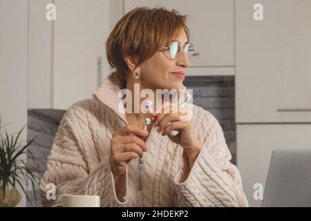 Bella donna boomer di mezza età in occhiali tiene le cuffie nelle sue mani e guarda lateralmente. Sfondo dell'interno grigio chiaro. Foto Stock