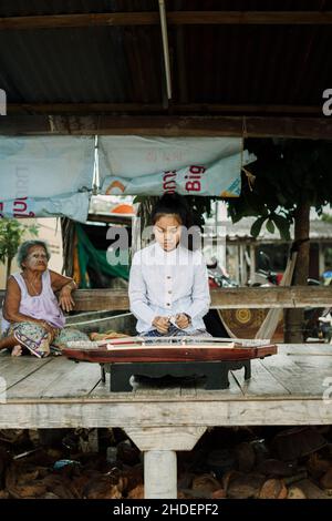 Una bella ragazza thailandese che suona Khim, il tradizionale strumento musicale thailandese vicino alla nonna all'aperto su pannello di legno. Concetto di tempo libero e hobby. Foto Stock