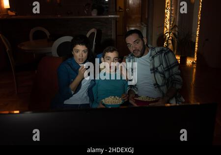 Famiglia di tre, padre, madre e figlio, guardando film horror in livingroom in buio. L'espressione del volto di Afrais e scioccata Foto Stock