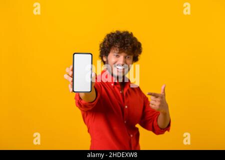 Ritratto di uomo bruna soddisfatto con capelli ricci puntati verso il cellulare e sorridendo alla fotocamera, consigliando gadget o applicazione mobile. Studio interno girato isolato su sfondo arancione Foto Stock