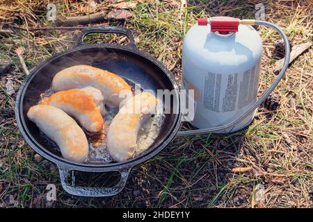 Salsicce in padella su bruciatore a gas turistico. Campeggio alimentare fare. Camper preparare il cibo nella foresta selvaggia in giorno di sole. Foto Stock