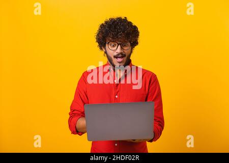 Ritratto di uomo sorpreso scioccato con la barba che tiene il laptop e guarda lo schermo con grandi occhi stupiti. Studio interno girato isolato su sfondo giallo Foto Stock