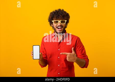 Felice uomo allegro bordato puntando dito su smartphone con schermo vuoto guardando la fotocamera con sorriso toothy, spazio libero per adv. Studio interno girato isolato su sfondo arancione Foto Stock