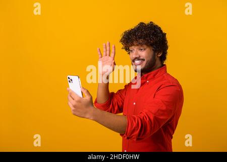 Uomo bearded positivo sorridente facendo selfie su smartphone fotocamera, blogger comunicazione, registrazione video per i follower in social network. Studio interno girato isolato su sfondo arancione Foto Stock