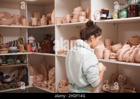 Vista posteriore di un vasaio femminile in piedi di fronte a ripiani con terracotta argilla ware Foto Stock
