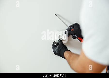 Primo piano delle mani di un operatore di sesso maschile, elettricista professionale in guanti di protezione utilizzando un cacciavite durante l'installazione di una nuova presa elettrica dopo Foto Stock