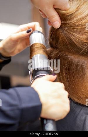 Mani di hairstylist femmina ricci i capelli cliente con un ferro da stiro in un parrucchiere, primo piano Foto Stock