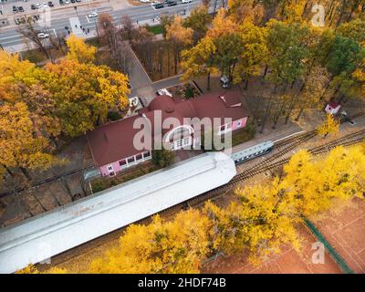 Guarda verso il basso sulla stazione ferroviaria sud dei bambini. Binario e piattaforma con treno in vividi alberi d'autunno gialli. Vista aerea sulla cima degli alberi sulla colorata Kha Foto Stock