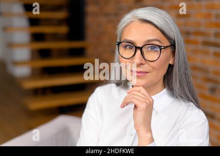 Ritratto di una donna di mezza età elegante ed elegante, che riposa il mento su una mano, sorridendo e guarda la macchina fotografica. Signora anziana dai capelli grigi seduta nel moderno appartamento e rilassante Foto Stock