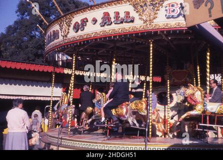 1960s, storico, persone, adulti e bambini, a cavallo su una giostra in una fiera, Inghilterra, Regno Unito. Un carosello, noto anche come giostra, è un giro nel parco divertimenti, un giro in piattaforma circolare in movimento, con posti fatti per assomiglia agli animali, in questo caso cavalli di legno decorati. Foto Stock