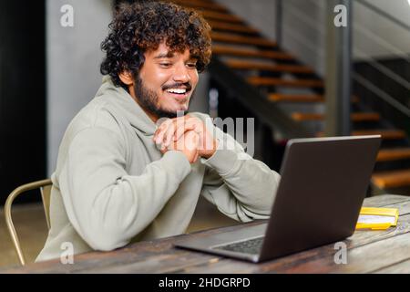 Vista laterale dell'uomo indiano ottimista che indossa una felpa con cappuccio seduta alla scrivania e un computer portatile a casa. Allegro studente di etnia maschile guarda lo schermo del computer con piacere, facendo videochiamata, guardando video Foto Stock
