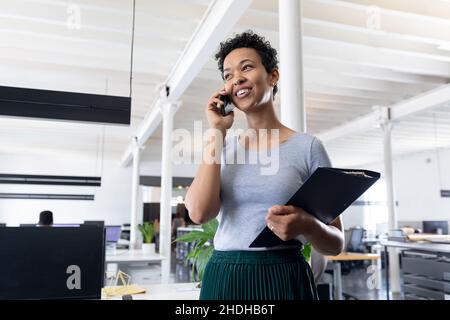 Sorridente donna d'affari biraciale che parla su smartphone in ufficio creativo Foto Stock