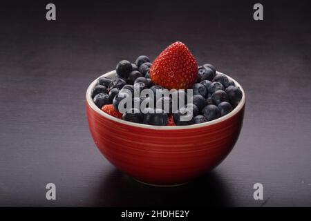 Ciotola rossa piena di mirtilli e fragole sul tavolo nero. Variopinto assortimento di succulenti frutta in attesa di essere consumata. Foto Stock