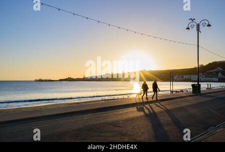 Camminatori del cane che camminano lungo il marciapiede alla baia di Swanage la mattina presto durante un'alba con il cielo blu e la stella del sole sopra la collina di Swanage vicino al punto di Peveril. Foto Stock