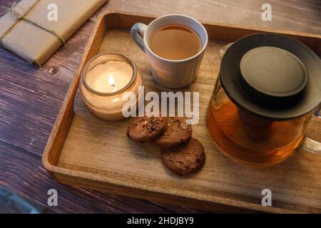 Concetto di coziness interno e domestico. Vista dall'alto. Candela che brucia in un vaso, una tazza di tè, una teiera con tè alle erbe e tre biscotti con scaglie di cioccolato di farinata d'avena Foto Stock