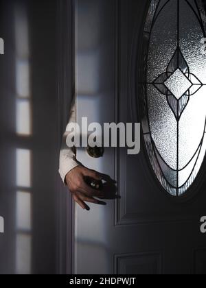 Mano dell'uomo che raggiunge per una maniglia della porta da dietro una porta di ingresso leggermente aperta in una casa buia con ombre spooky di notte. Furto, crimine, omicide Foto Stock