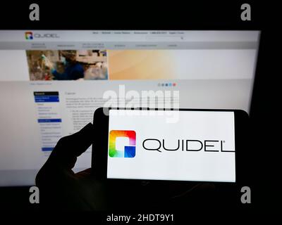 Persona che tiene il cellulare con il logo della società americana di diagnostica Quidel Corporation sullo schermo di fronte alla pagina web. Mettere a fuoco sul display del telefono. Foto Stock