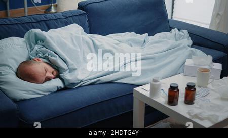 Primo piano di adulto con sintomi influenzali che si trovano sul divano per curare il raffreddore e la malattia da virus. Uomo malato che dorme e che ha trattamento di prescrizione, bottiglie di pillole, capsule e termometro sul tavolo Foto Stock