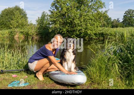 Peize, Olanda 16 luglio 2021: Una donna abbronzata in t-shirt e shorts. Prepara la tavola sup per la vela. Il suo cane pastore tedesco è seduto Foto Stock