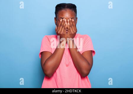 Ritratto di spaventato stressato ragazza afroamericana coprendo il volto con le mani durante foto shotting avendo ansia sociale in piedi in studio con sfondo blu. Terrificato modello introvert Foto Stock