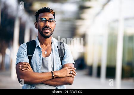 Ritratto di uomo nero sorridente con occhiali e zaino con braccia incrociate. Foto Stock