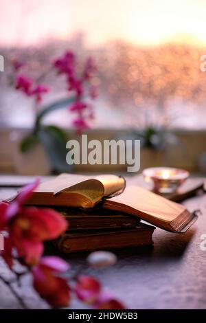 Inverno con candela su pila di vecchi libri. Finestra al tramonto con bagliore arancione, orchidea rosa e fucsia e fiori di magnolia. Candele aromatiche, tè Foto Stock
