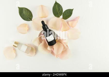 due flaconi cosmetici con pipetta e siero per la cura della pelle del viso si trovano su petali di rosa su sfondo bianco. Mockup senza marchio Foto Stock